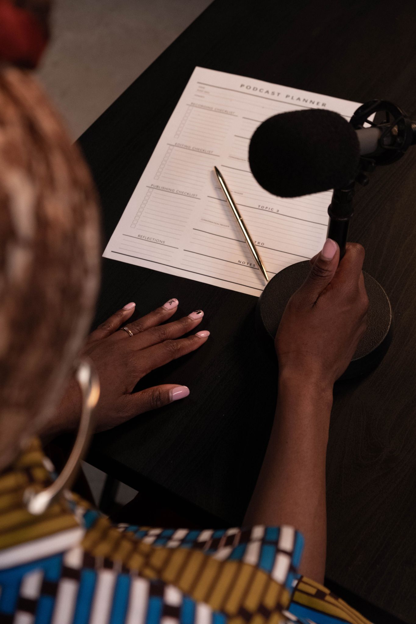 Eine Draufsicht auf eine Person mit einem Mikrofon und einem Podcast-Planer auf einem Schreibtisch (Quelle: pexels|cottonbro studios)